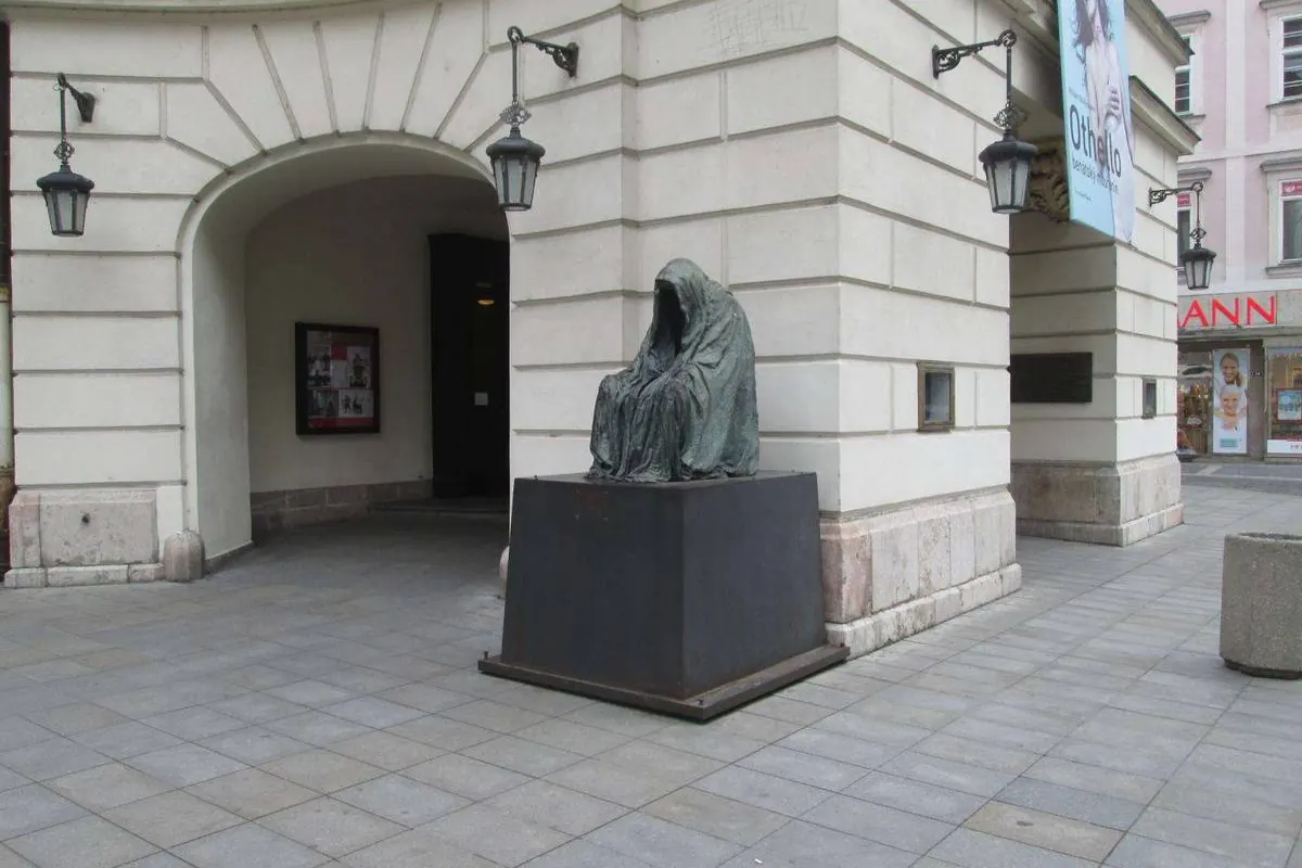 Eine Bronzestatue einer vermummten, gesichtslosen Figur auf dunklem Sockel unter einer bogigen weißen Wand, vor dem Ständetheater.