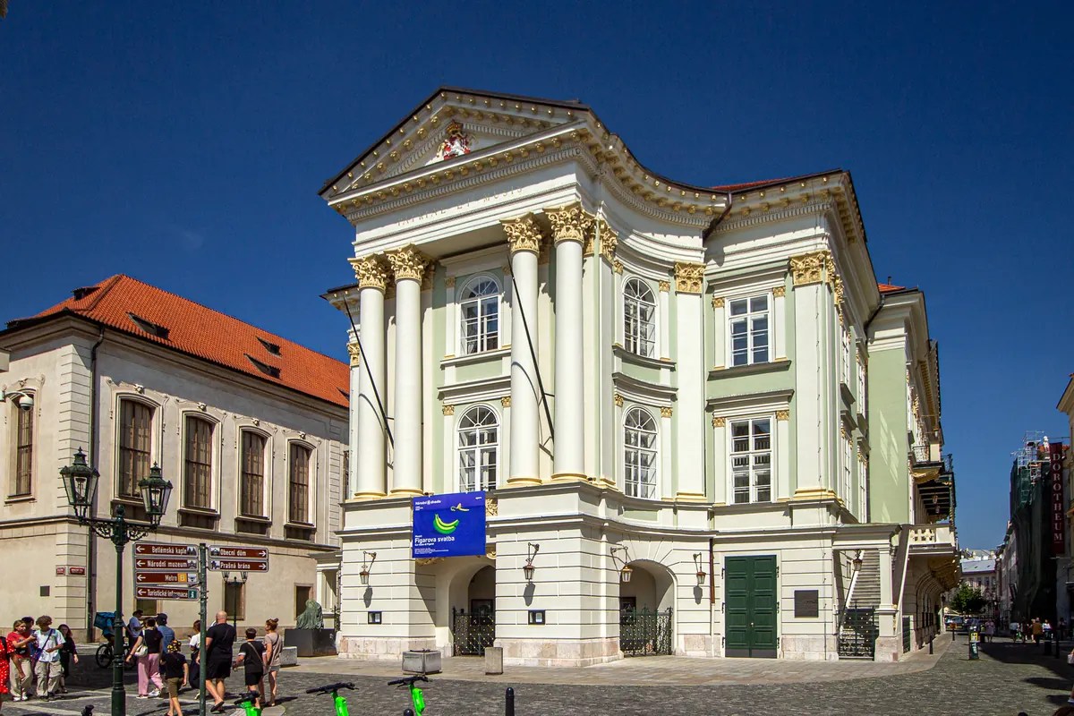 Die cremefarbene und hellgrüne neoklassizistische Fassade des Ständetheaters mit weißen Säulen unter tiefblauem Himmel.